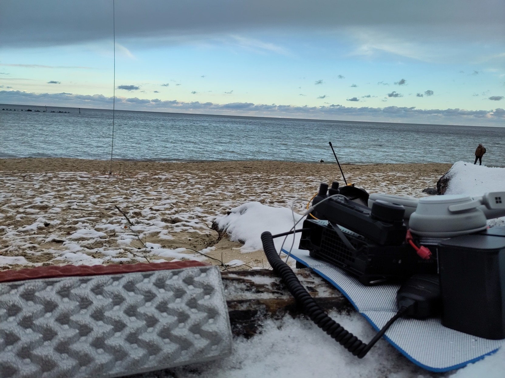 Im Hintergrund sieht man das Meer und einen Sandstrand, leicht mit Schnee bedeckt, im Vordergrund eine Sitzunterlage, Dunkgerät, Kopfhörer Akk und Handmikrofon. Eine Antenne ist als vertikaler Strich zu erkennen